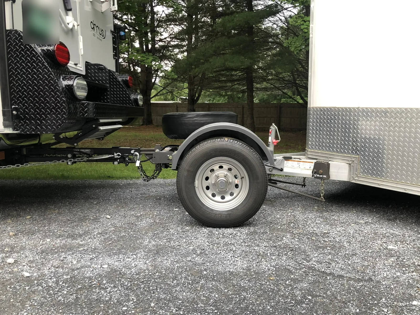 Close-up of a Trailer Toad hitched between the back of a truck camper and the front of a white enclosed trailer. The weight distribution hitch runs from the camper to the Toad, the Toad sits on one wheel under a fender, and the trailer's coupler sits on the ball at the rear of the Toad.