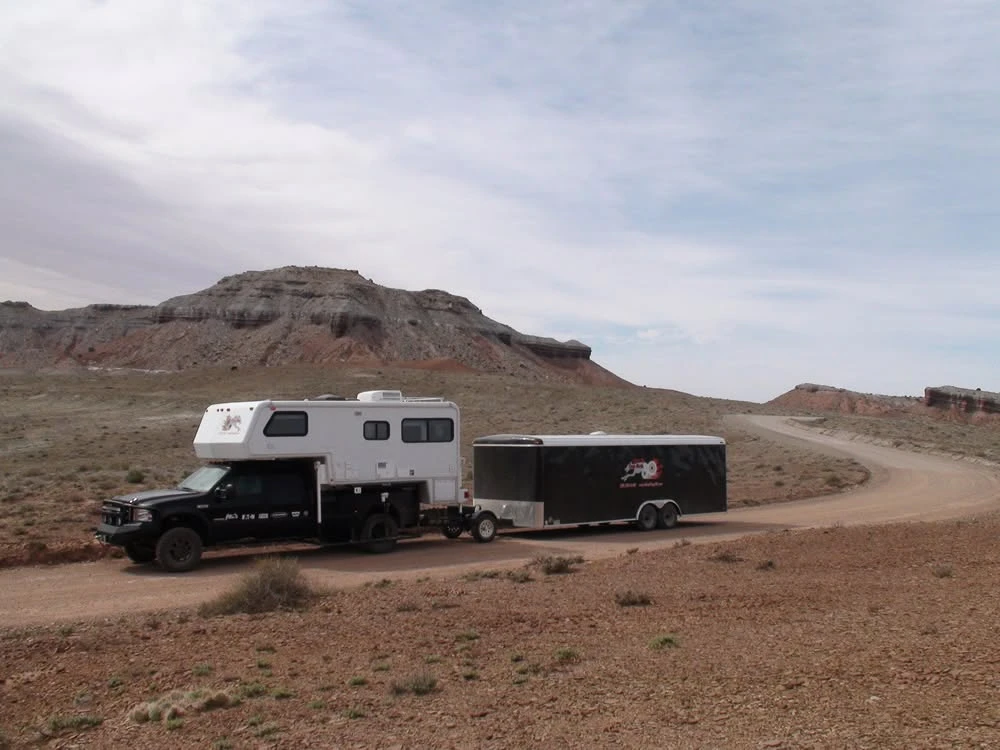 A black pickup with a truck camper towing a black enclosed car hauler on a desert dirt road. A Trailer Toad rides between the camper and the hauler, on its own single wheel.