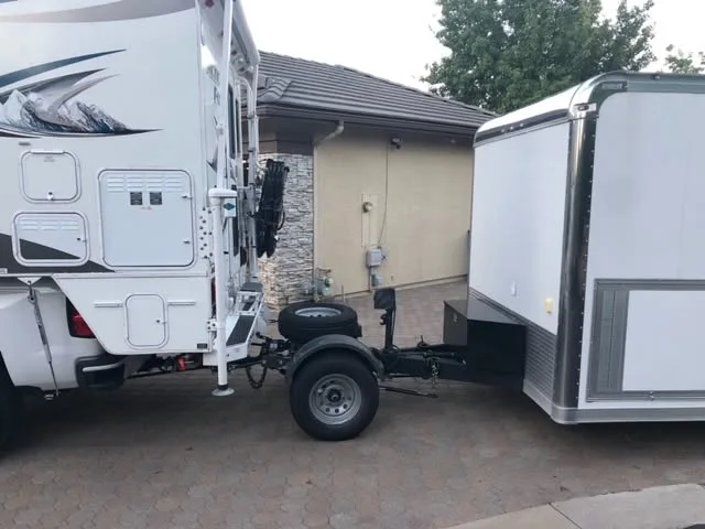 A Trailer Toad with a spare tire on top, on one wheel between the back of a white truck camper and the front of a white enclosed trailer, parked on a paver driveway.
